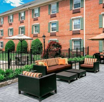 Sonesta Select Tinton Falls Eatontown’s patio is furnished with shaded tables and chairs, in addition to sofa and armchair seating, with greenery nearby.