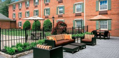 Sonesta Select Tinton Falls Eatontown’s patio is furnished with shaded tables and chairs, in addition to sofa and armchair seating, with greenery nearby.