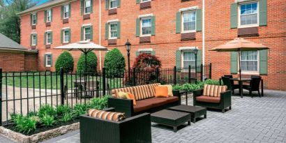 Sonesta Select Tinton Falls Eatontown’s patio is furnished with shaded tables and chairs, in addition to sofa and armchair seating, with greenery nearby.