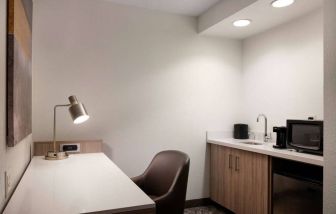 Guest room workspace in Sonesta Select Seattle Renton, including desk, chair, and lamp, with kitchenette nearby.