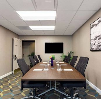 Hotel meeting room, featuring long wooden table, eight swivel chairs, and a large, widescreen TV on the wall.