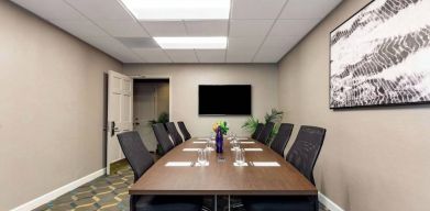 Hotel meeting room, featuring long wooden table, eight swivel chairs, and a large, widescreen TV on the wall.