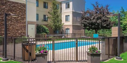 Sonesta ES Suites Andover Boston’s outdoor pool is ringed by a fence, with a backdrop of the hotel and numerous trees.