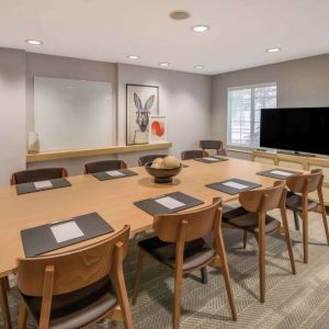 Hotel meeting room, with long wooden table and seating for nine, art and a whiteboard on the wall, and a large television. 