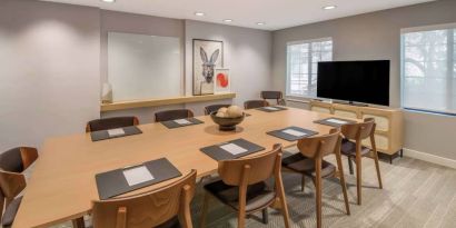 Hotel meeting room, with long wooden table and seating for nine, art and a whiteboard on the wall, and a large television. 