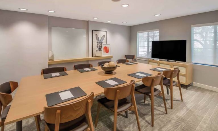 Hotel meeting room, with long wooden table and seating for nine, art and a whiteboard on the wall, and a large television. 