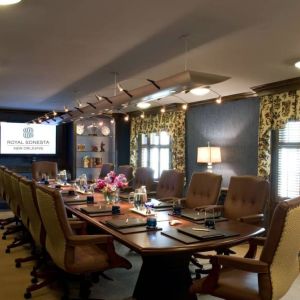 Meeting room in Royal Sonesta New Orleans, furnished with long wooden table, 14 swivel chairs, a widescreen TV, and two windows.