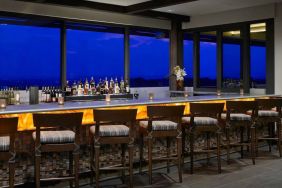 The hotel bar has traditional tall bar stools, a hard floor, a wide array of beverages, and large windows.