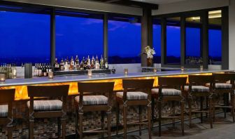 The hotel bar has traditional tall bar stools, a hard floor, a wide array of beverages, and large windows.