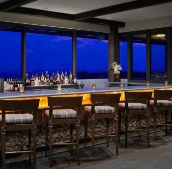 The hotel bar has traditional tall bar stools, a hard floor, a wide array of beverages, and large windows.