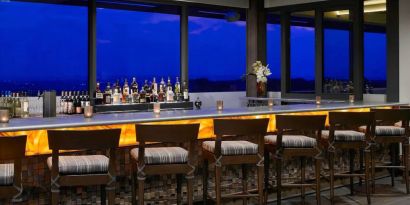 The hotel bar has traditional tall bar stools, a hard floor, a wide array of beverages, and large windows.