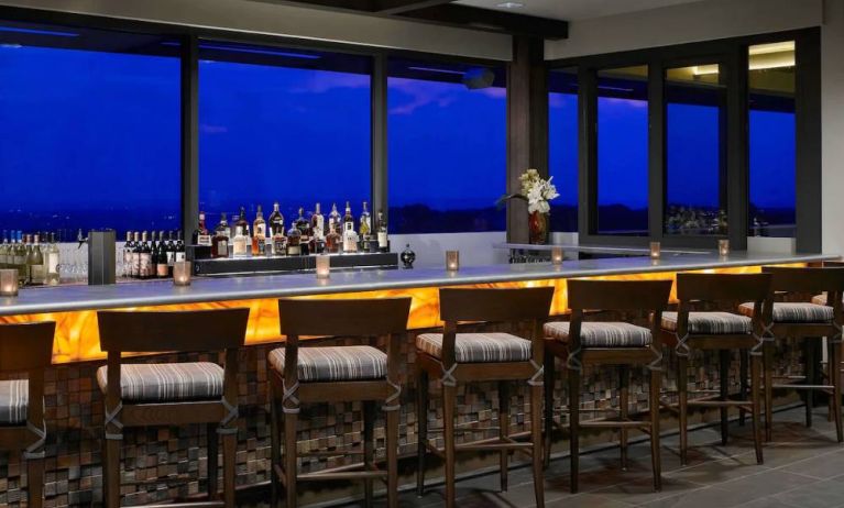 The hotel bar has traditional tall bar stools, a hard floor, a wide array of beverages, and large windows.