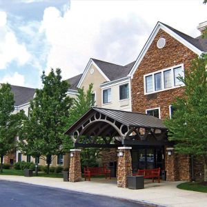 The hotel’s exterior features a covered entranceway with a pair of benches, flanked by numerous trees and bushes.