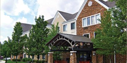 The hotel’s exterior features a covered entranceway with a pair of benches, flanked by numerous trees and bushes.