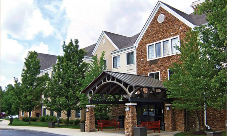 The hotel’s exterior features a covered entranceway with a pair of benches, flanked by numerous trees and bushes.