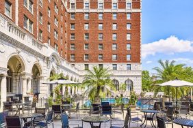 The hotel patio features tables and chairs, some shaded, overlooking the outdoor pool.