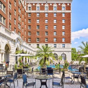 The hotel patio features tables and chairs, some shaded, overlooking the outdoor pool.