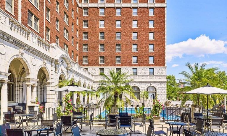 The hotel patio features tables and chairs, some shaded, overlooking the outdoor pool.