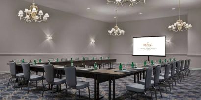 Meeting room in The Chase Park Plaza Royal Sonesta St. Louis, featuring long tables arranged in a U-shape, quartet of chandeliers, seating for over a dozen, and a projector screen.