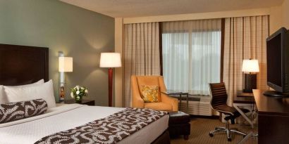 Double bed guest room in Sonesta Atlanta Airport North, featuring TV, window, armchair, and workspace desk and chair.