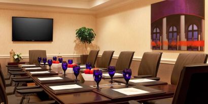 Meeting room in Sonesta Atlanta Airport North, furnished with long wooden table, a dozen chairs, and art and a widescreen television on the walls.