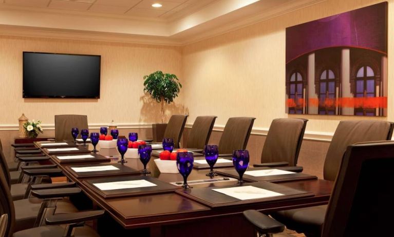 Meeting room in Sonesta Atlanta Airport North, furnished with long wooden table, a dozen chairs, and art and a widescreen television on the walls.