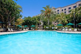 The outdoor pool at Sonesta ES Suites Anaheim Resort Area features nearby sun loungers and shaded tables and chairs, with numerous trees close by.