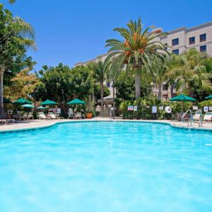 The outdoor pool at Sonesta ES Suites Anaheim Resort Area features nearby sun loungers and shaded tables and chairs, with numerous trees close by.