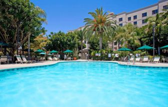 The outdoor pool at Sonesta ES Suites Anaheim Resort Area features nearby sun loungers and shaded tables and chairs, with numerous trees close by.