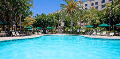 The outdoor pool at Sonesta ES Suites Anaheim Resort Area features nearby sun loungers and shaded tables and chairs, with numerous trees close by.