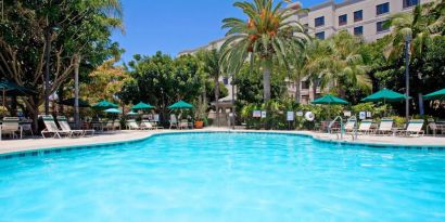 The outdoor pool at Sonesta ES Suites Anaheim Resort Area features nearby sun loungers and shaded tables and chairs, with numerous trees close by.