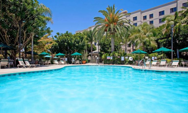 The outdoor pool at Sonesta ES Suites Anaheim Resort Area features nearby sun loungers and shaded tables and chairs, with numerous trees close by.