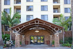 The hotel's exterior features a covered entrance, with potted plants and large trees and other greenery to either side.