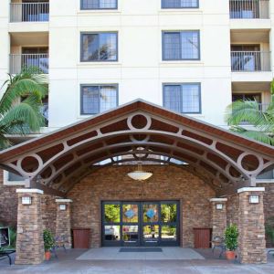 The hotel's exterior features a covered entrance, with potted plants and large trees and other greenery to either side.