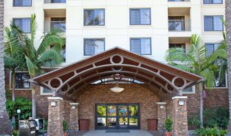 The hotel's exterior features a covered entrance, with potted plants and large trees and other greenery to either side.