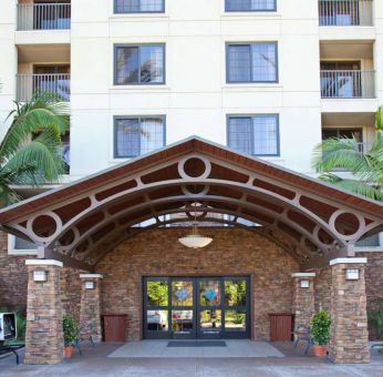 The hotel's exterior features a covered entrance, with potted plants and large trees and other greenery to either side.