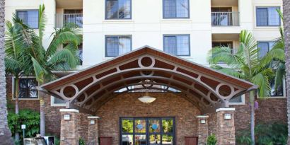 The hotel's exterior features a covered entrance, with potted plants and large trees and other greenery to either side.