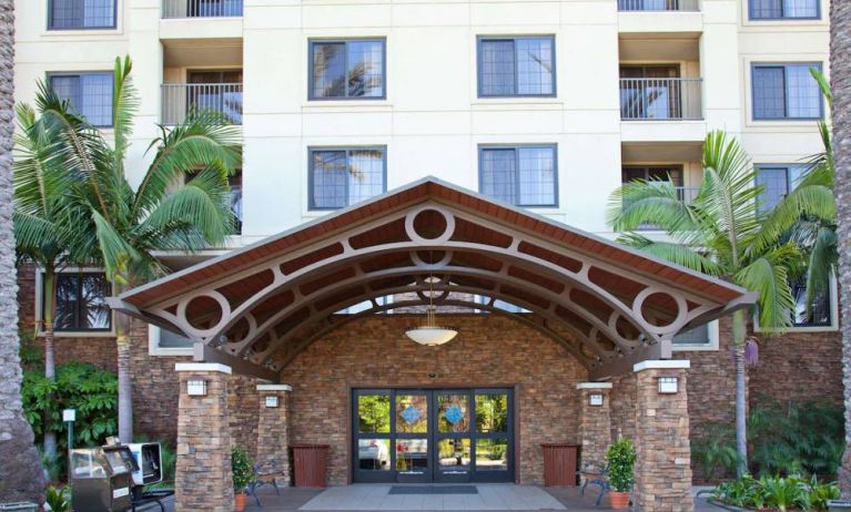 The hotel's exterior features a covered entrance, with potted plants and large trees and other greenery to either side.