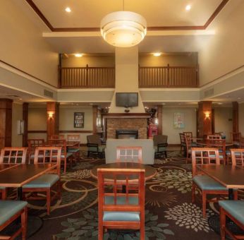 The hotel breakfast area has a high ceiling, wide range of table sizes, carpeted floor, and a wall-mounted TV.