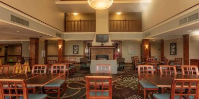 The hotel breakfast area has a high ceiling, wide range of table sizes, carpeted floor, and a wall-mounted TV.