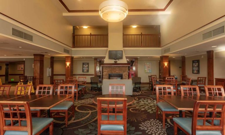 The hotel breakfast area has a high ceiling, wide range of table sizes, carpeted floor, and a wall-mounted TV.