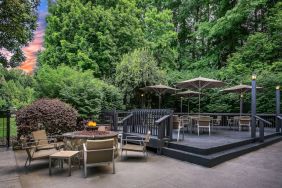 Sonesta Charlotte Executive Park’s patio includes shaded tables and chairs, and a fire pit surrounded by chairs and coffee tables, with abundant greenery nearby.