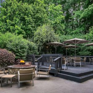 Sonesta Charlotte Executive Park’s patio includes shaded tables and chairs, and a fire pit surrounded by chairs and coffee tables, with abundant greenery nearby.