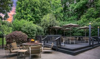 Sonesta Charlotte Executive Park’s patio includes shaded tables and chairs, and a fire pit surrounded by chairs and coffee tables, with abundant greenery nearby.