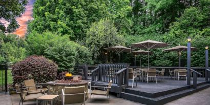 Sonesta Charlotte Executive Park’s patio includes shaded tables and chairs, and a fire pit surrounded by chairs and coffee tables, with abundant greenery nearby.