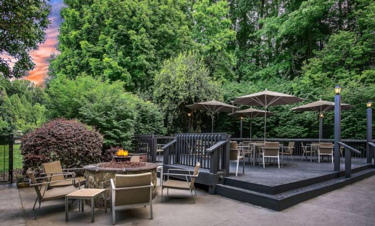 Sonesta Charlotte Executive Park’s patio includes shaded tables and chairs, and a fire pit surrounded by chairs and coffee tables, with abundant greenery nearby.