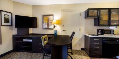 Sonesta Simply Suites St Louis Earth City guest room workspace, furnished with desk and chairs, with TV and kitchen nearby.