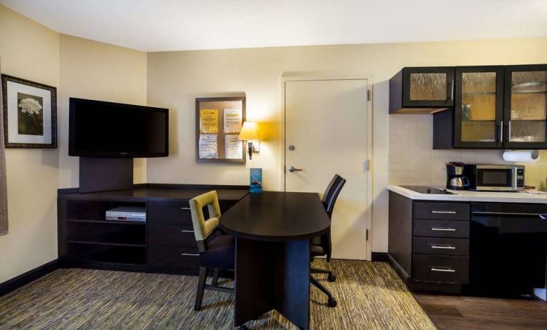 Sonesta Simply Suites St Louis Earth City guest room workspace, furnished with desk and chairs, with TV and kitchen nearby.