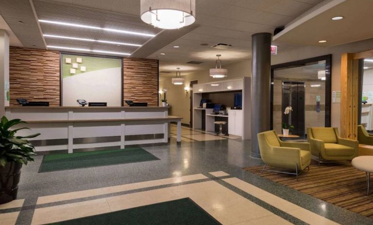 The hotel lobby has yellow armchairs, potted plants, and a business center workstation, in addition to the reception desk.