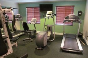 The hotel fitness center has two windows fitted with blinds, a wall-mounted TV, and  assorted exercise machines.